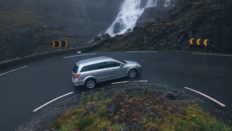 Car-driving-uphill,-moving-through-a-sharp-turn,-beautiful-scenic-view-of-mountain-waterfalls-reveals-as-car-drives-out-of-frame