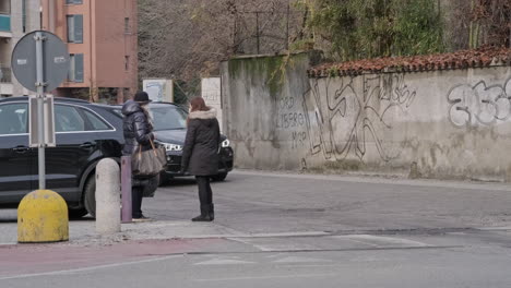 Dos-Mujeres-Enmascaradas-Hablando-En-El-Lado-De-La-Calle-Con-Autos-Pasando-En-Arcore,-Norte-De-Italia