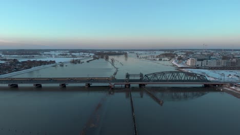 Vorwärtsbewegung-Sonnenuntergangsantenne-Zeigt-überflutete-Auen-Des-Flusses-Ijssel,-Vorbei-An-Der-Niederländischen-Hanseatischen-Mittelalterlichen-Turmstadt-Zutphen,-Mit-Einem-Zug,-Der-über-Die-Stählerne-Zugbrücke-Vorbeifährt