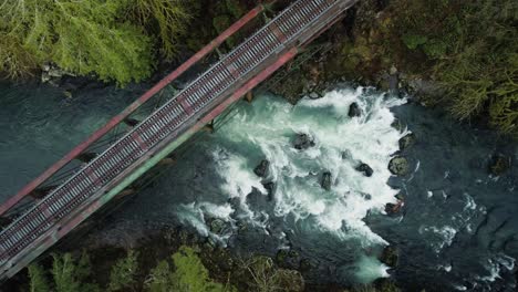 Lewis-River-flows-downstream-under-steel-railroad-bridge-through-rapids,-aerial-static-shot