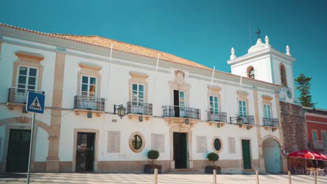Portugal-Algarve-Loule-Typisches-Quadratisches-Rathaus-Mit-Vorbeifahrenden-Fußgängern,-Autos-Und-Motorrädern-Am-Morgen-Mit-Sonnenschein-4k