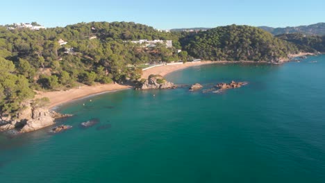 Aerial,-drone-shot,-over-rocky-coast-and-turquoise-sea,-towards-Lloret-de-Mar-beach,-Santa-Cristina,-Cala-Boadella-,-in-Costa-Brava,-Catalonia,-Spain