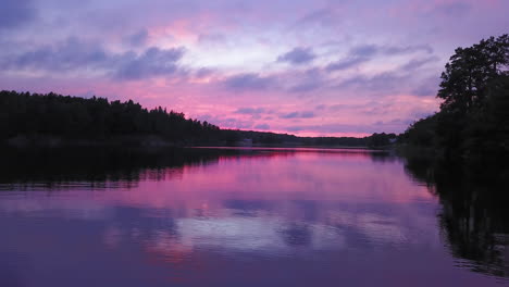 Aerial-drone-shot-close-to-the-water,-above-a-lake-and-towards-the-forest,-a-purple-sky,-at-a-colorful-sunset-or-dusk,-at-Albysjon,-Tyreso,-Sweden