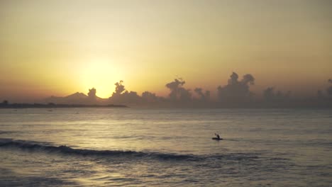 Ein-Mann-Auf-Einem-Kleinen-Traditionellen-Boot,-Das-Bei-Sonnenuntergang-Oder-Sonnenaufgang-Mit-Dramatischen-Wolken-In-Bali,-Indonesien,-In-Zeitlupe-In-Richtung-Meer-Segelt