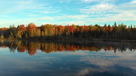 Aerial-footage-sliding-left-across-glassy-pond-with-forested-shore-in-fall-colors