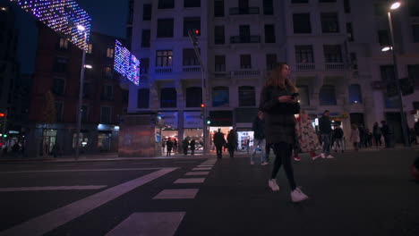 Low-panning-shot-across-front-of-vehicle---headlights-to-reveal-people-crossing-road---walking-sidewalk-in-Madrid-retail-area-illuminated-at-night