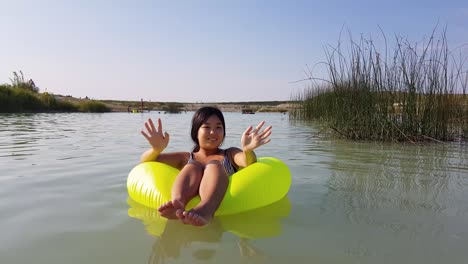 Girl-sitting-in-yellow-inflatable-tire-floating-in-lake,-looking-and-waving-at-camera-during-beautiful-summer-day