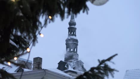 Church-tower-in-Tallinn-old-town-seen-between-the-branches-of-a-christmas-tree-decorated-with-lights,-and-some-snow-covering-the-roofs-in-the-evening