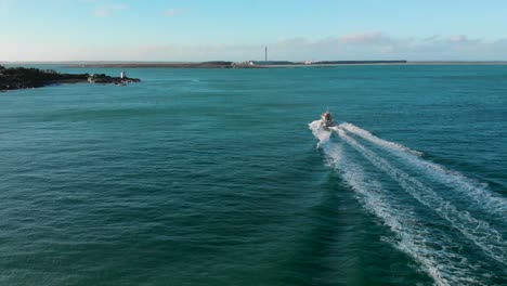Following-commercial-boat-on-blue-ocean-beautiful-sunny-day-with-lighthouse-and-coast-in-the-background---Bluff,-New-Zealand---Aerial-Drone