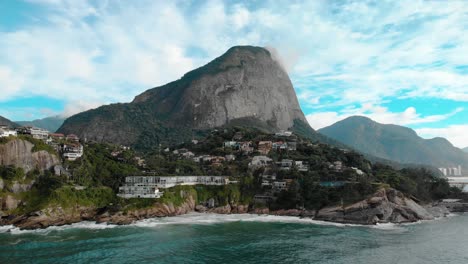 Aerial-approach-of-the-beautiful-picturesque-cliff-rocks-of-Joatinga-beach-in-Rio-de-Janeiro-with-the-Gavea-mountain-towering-behind-it-and-the-green-ocean-waves-rolling-in