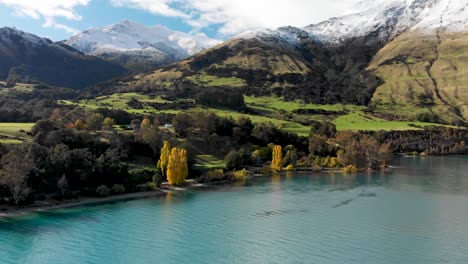 SLOWMO---Flying-over-beautiful-blue-Lake-Wakatipu,-Queenstown,-New-Zealand-with-mountains-fresh-snow,-clouds,-blue-sky-autumn-trees-in-the-background---Aerial-Drone