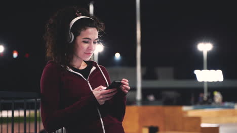 Smiling-caucasian-sportswoman-listening-music-with-bluetooth-headphones-and-texting-on-her-mobile-phone-while-taking-a-break-during-her-training-session-at-night-in-the-park
