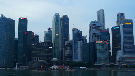 Singapur---Circa-2016time-Lapse-De-Rascacielos-Iluminados-Frente-Al-Mar-En-Singapur-Durante-El-Crepúsculo-De-La-Tarde,-Barcos-En-El-Río