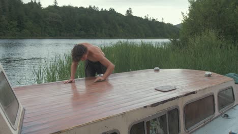 Fitter-Junger-Mann-Oben-Ohne-Schleifen-Der-Holzdachplanke-Eines-Alten-Holzbootes