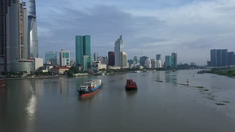 Drohnenaufnahme-Eines-Frachtschiffs-Und-Eines-Kleineren-Schiffes,-Die-Schiffscontainer-Entlang-Des-Saigon-Flusses-Transportieren,-Mit-Blick-Auf-Das-Finanz--Und-Stadtzentrum