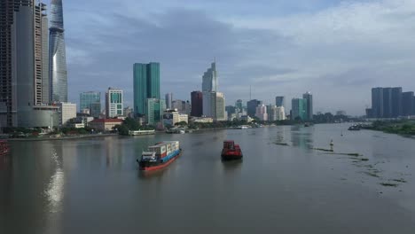 drone-shot-of-A-cargo-ship-and-smaller-vessel-are-transporting-shipping-containers-along-the-Saigon-river-with-the-financial-and-city-center-in-full-view