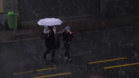 Mother-and-sunning-crossing-a-street-during-a-hail-storm,-protecting-themselves-with-an-umbrella-and-a-hood