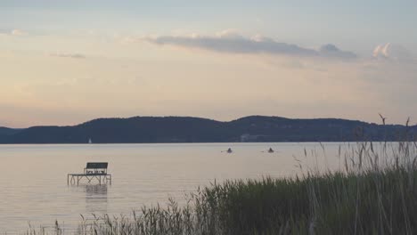 Zwei-Personen-Paddeln-In-Booten-Auf-Einem-Ruhigen-See-Bei-Sonnenuntergang-Mit-Einem-Segelboot-Und-Hügeln-Im-Hintergrund