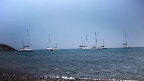 Freihandaufnahme-Von-Acht-Segelyachten,-Die-Am-Roten-Strand-In-Santorini,-Griechenland,-Versammelt-Sind,-Und-Eines-Mannes,-Der-Schwimmt