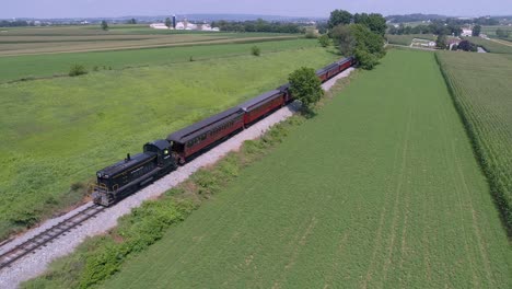 Una-Vista-Aérea-De-Una-Locomotora-Diésel-Tirando-De-Coches-De-Pasajeros-Antiguos-A-Través-De-La-Campiña-Amish