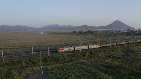 Locomotoras-De-Trenes-Rusos-Tirando-De-Un-Largo-Vagón-De-Carbón-Vacío-Cargando-A-Través-De-Campos-Verdes-Con-Montañas-En-El-Fondo-De-La-Puesta-Del-Sol,-Federación-Rusa