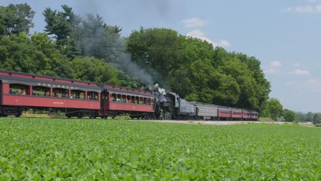 Eine-Dampflokomotive-Aus-Dem-Jahr-1910-Mit-Personenzug-Fährt-In-Einen-Bahnhof-Ein-Und-Bläst-Schwarzen-Rauch-Durch-Die-Amish-Landschaft,-Während-An-Einem-Sonnigen-Sommertag-Eine-Zweite-Dampflokomotive-Vorbeifährt