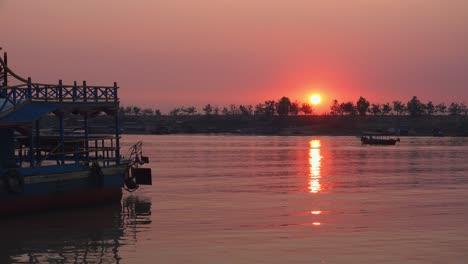 Zeitraffer-Einer-Wunderschönen-Orangefarbenen-Sonne,-Die-Im-Hintergrund-über-Dem-Horizont-Des-Anderen-Ufers-Des-Sees-Untergeht,-Mit-Einem-Boot,-Das-Zum-Parken-Kommt,-Und-Einem-Geparkten-Blauen-Touristenboot,-Das-Im-Wasser-Auf-Und-Ab-Schaukelt