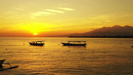 Siluetas-De-Barcos-Flotando-En-La-Bahía-Tranquila-Al-Atardecer-Con-Un-Cielo-Naranja-Sobre-El-Horizonte-De-La-Montaña-Que-Se-Refleja-En-La-Superficie-Del-Mar,-Bali