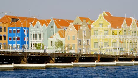 Gente-Caminando-En-El-Puente-De-La-Reina-Emma-Con-Edificios-Coloridos-En-El-Fondo-En-Un-Día-Soleado-En-Punda,-Curacao