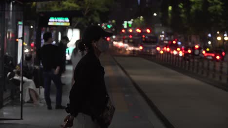 Korean-woman-wearing-face-mask-stands-by-bus-stop-in-downtown-city-at-night,-covid-19,-Seoul,-South-Korea
