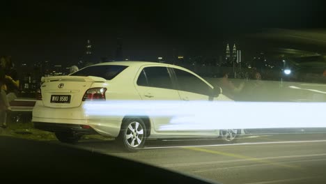 Weißes-Auto-Am-Straßenrand-In-Kuala-Lumpur,-Malaysia-Geparkt,-Mit-Vorbeifahrenden-Autos-Auf-Der-Straße-In-Der-Nacht---Zeitraffer