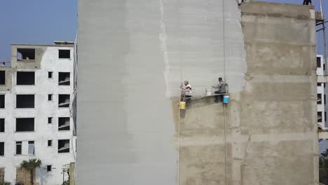 Trabajadores-De-La-Construcción-Colgados-En-El-Costado-De-Un-Edificio-Aplicando-Acabado-De-Yeso-De-Hormigón,-órbita-Aérea-Alrededor-Del-Tiro