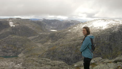 Eine-Frau-Lächelt,-Während-Sie-Von-Einem-Berg-In-Norwegen-Aus-Einen-Atemberaubenden-Blick-Auf-Eine-Felsige,-Hochgelegene-Landschaft-Genießt,-Zeitlupe