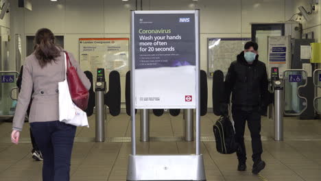 People-in-protective-surgical-face-masks-enter-and-exit-an-underground-station-and-pass-an-NHS-health-sign-waring-the-to-public-to-wash-hands-for-twenty-seconds-during-the-Coronavirus-outbreak