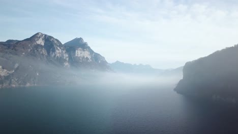 Panoramic-shot-of-low-hanging-fog-between-mountain-ranges-above-the-water-surface-of-a-crystal-blue-mountain-lake