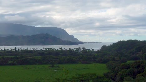 HD-Hawaii-Kauai-slow-motion-static-wide-shot-of-a-grassy-field-with-a-cove-and-mountainous-coastline-in-distance-with-mostly-cloudy-sky