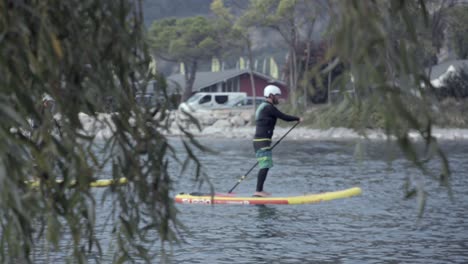 Watching-man-pass-on-paddle-board-through-low-branches,-focus-pull