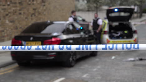 Pull-focus-on-a-blue-and-white-“police-line-do-not-cross”-tape-flutters-in-the-breeze-as-two-police-officers-guard-a-black-BMW-car-at-the-scene-of-a-knife-crime-incident
