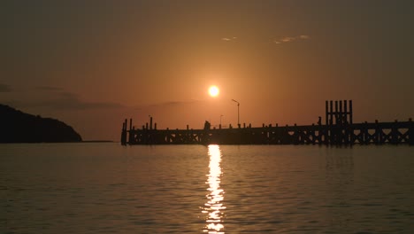 Warm-orange-sun-above-sea-with-reflection-on-water-surface-and-pier-silhouette-during-sunset