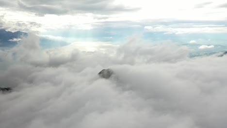 Drone-shot-moving-forwards,-revealing-a-mountain-top-barely-peeking-through-the-clouds-that-are-moving-fast,-passing-by