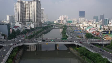 Drone-flight-hovering-over-Ben-Nghe-canal-featuring-Saigon,-Vietnam-City-skyline,-Calmette-and-Ong-Lanh-bridges-and-foggy-morning-day-time-traffic