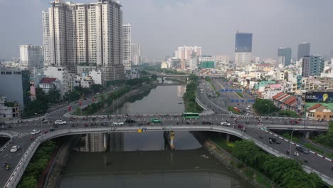 Vuelo-De-Drones-Sobrevolando-El-Canal-Ben-Nghe-Con-Saigon,-El-Horizonte-De-La-Ciudad-De-Vietnam,-Los-Puentes-Calmette-Y-Ong-Lanh-Y-El-Tráfico-Matutino-Brumoso