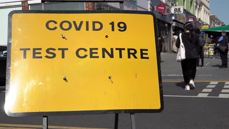 A-woman-in-a-protective-face-mask-walks-past-a-yellow-Covid-19-Test-Centre-street-sign-on-a-high-street