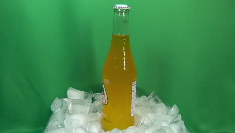 1-2-Botellas-De-Bebida-De-Mango-Carbonatado-Que-Giran-360-Grados-Sobre-Hielo-Apilado-Frente-A-Una-Pantalla-Verde