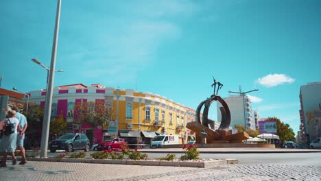 Portugal-Algarve-Loule-Street-Fuente-Plaza-Rotonda-Con-Pareja-De-Peatones,-Coches-Pasando-Por-La-Mañana-Con-Sol-4k