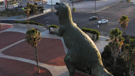 Aerial-view-of-a-T-Rex,-one-of-the-Cabazon-Dinosaurs,-World's-Biggest-Dinosaurs,-California,-next-to-palm-trees