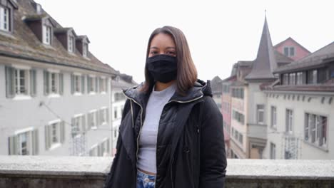 Slow-motion-shot-of-attractive-female-model-with-respirator-mask-against-virus-infection-standing-in-front-of-city-of-Rapperswil,-Switzerland