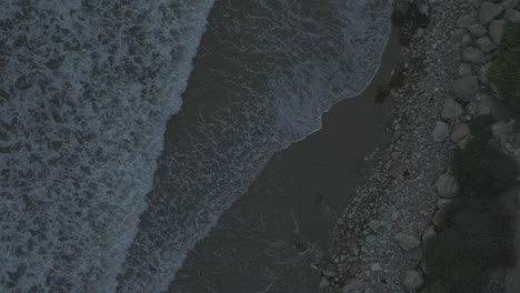 Top-down-view-of-ocean-waves-crashing-against-an-rocky-sand-beach-from-above