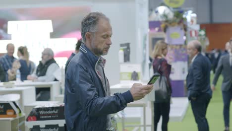 Man-standing-around,-looking-down-at-phone,-busy-conference,-people-in-background