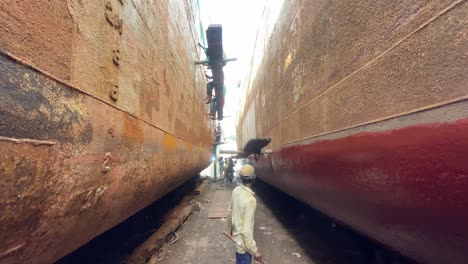 Adolescente-Pintando-Pintura-De-Casco-De-Barco-De-Pie-Entre-Dos-Viejos-Barcos-Oxidados-Con-Gente-Trabajadora-En-El-Fondo-En-Dhaka,-Bangladesh